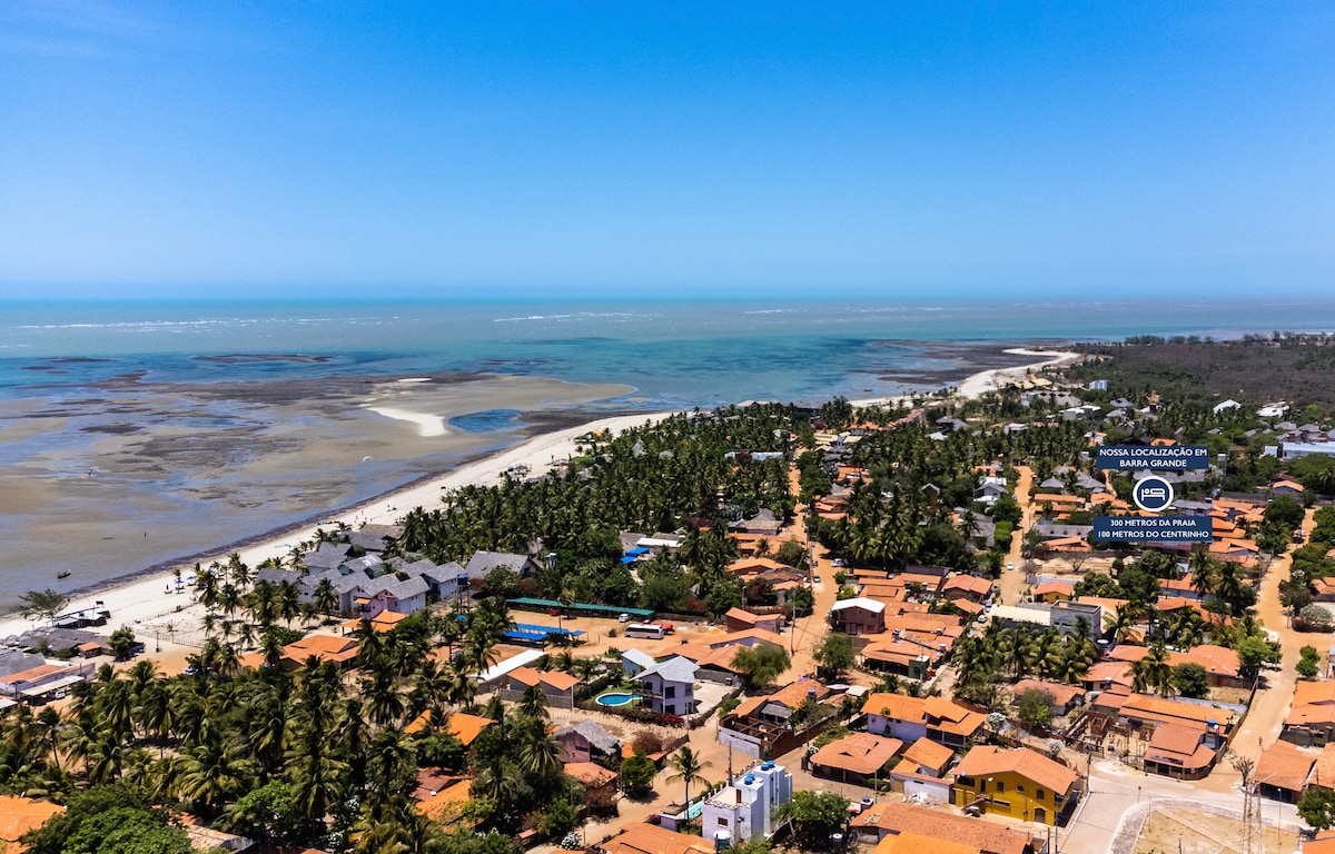An aerial view reveals a coastal village bordered by a sandy beach and turquoise waters. Homes with terracotta roofs nestle among lush greenery, while the shoreline is lined with palm trees. The scene is tranquility, showcasing a blend of natural beauty and community charm.