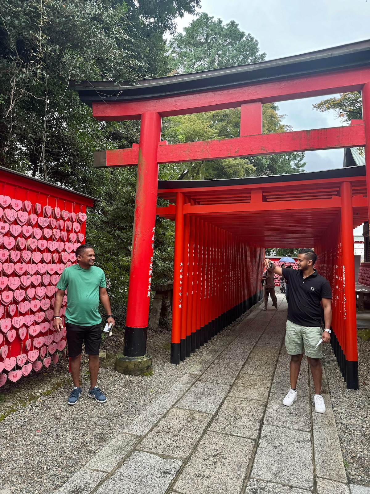 A vibrant red torii gate stands prominently at the entrance to a shrine pathway. On either side, walls adorned with wooden heart-shaped plaques create a contrasting backdrop. Visitors can be seen engaged with the surroundings, enhancing the cultural atmosphere of the location.