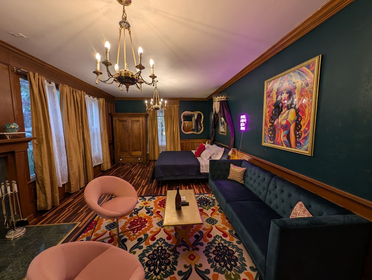 A spacious living area features bold blue walls accented by elegant wooden trim. A vibrant area rug lies beneath a coffee table, with two pink chairs and a plush sofa inviting relaxation. Large windows draped with golden curtains allow natural light to brighten the room.