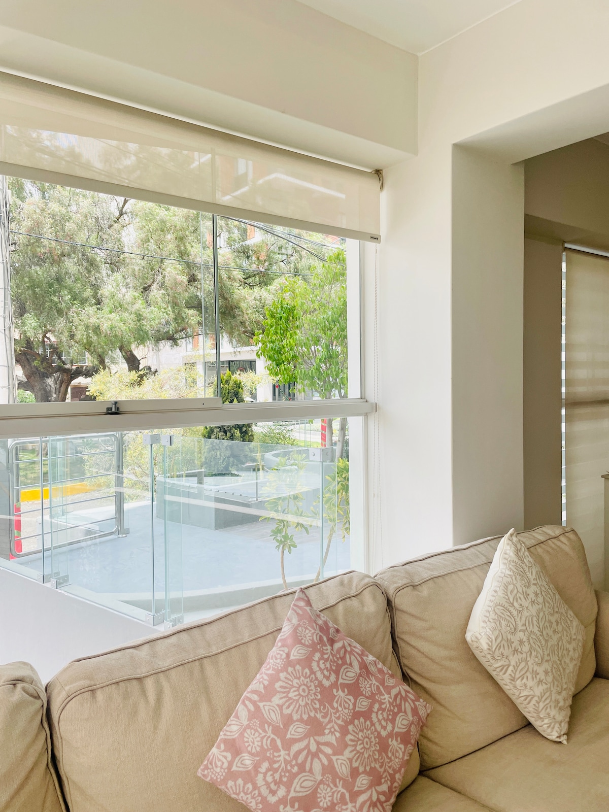 A comfortable seating area with a light-toned couch and decorative pillows is positioned beside a large window. Natural light filters through the blinds, illuminating the greenery outside, including trees and landscaped areas visible from the interior.