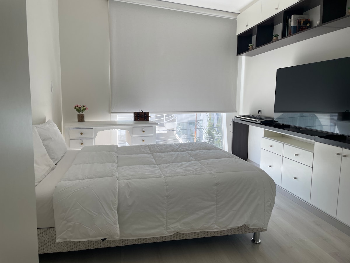 A well-lit bedroom features a spacious bed covered with a white comforter. A simple desk with a plant is positioned near the window, while a modern media unit and shelves are arranged along the wall opposite the bed, providing both functionality and storage.