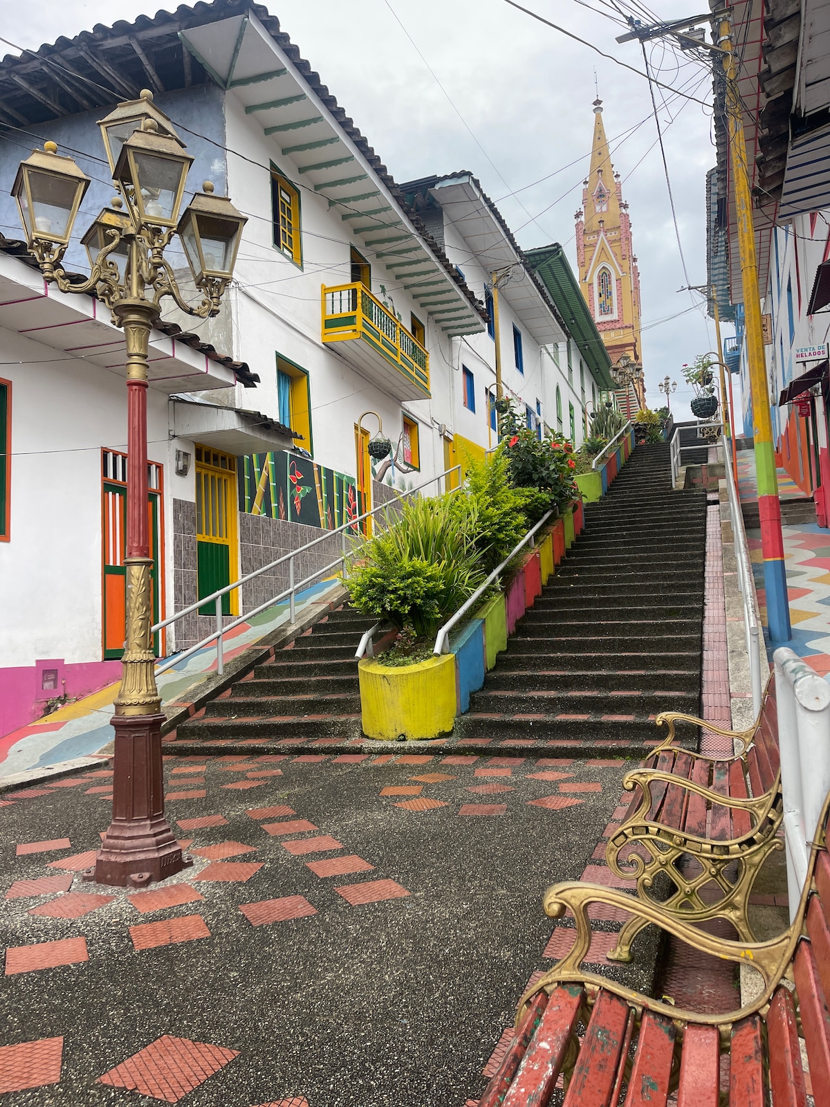 A vibrant street scene is depicted, showcasing a steep staircase lined with colorful planters and benches. Charming white buildings feature yellow and red accents, while a tall church spire rises in the background, contributing to the serene, inviting atmosphere of the area.