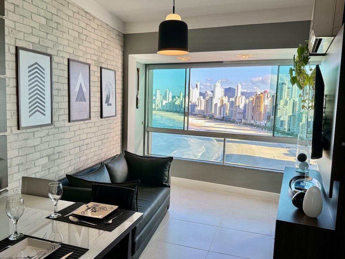 A stylish living space is showcased with a sleek black sofa and a dining table set for two. Large windows provide a panoramic view of the beach and city skyline, allowing natural light to illuminate the modern décor.