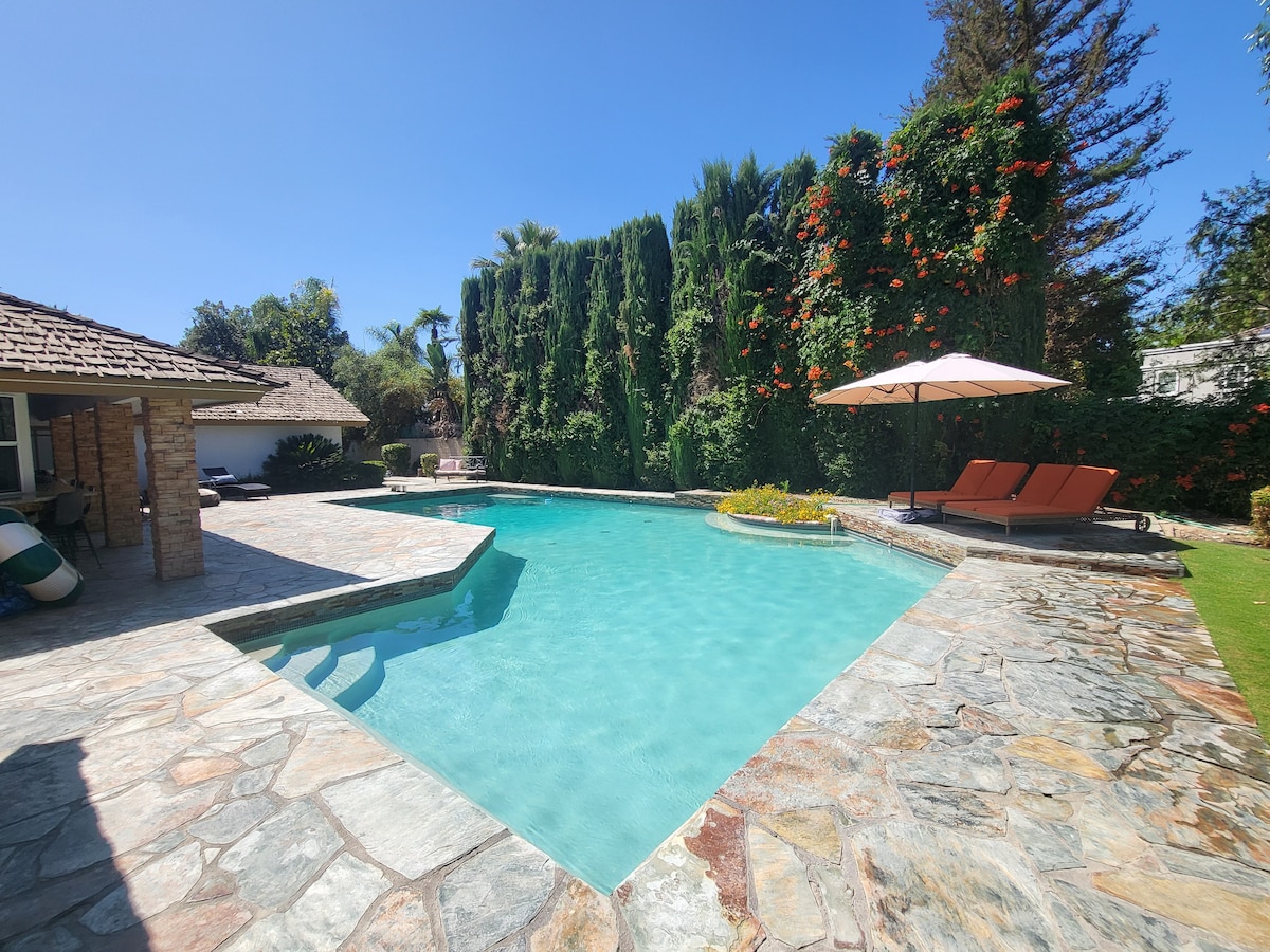 A spacious backyard features a sparkling swimming pool surrounded by natural stone decking. Two lounge chairs are positioned under an umbrella, offering a shaded relaxation area. Lush greenery and vibrant flowering plants create a serene environment, enhancing the outdoor space.