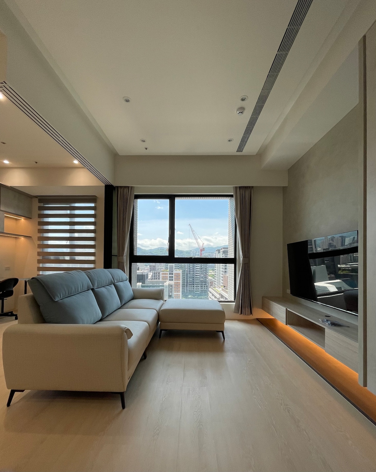 A modern living area features a light-colored sectional sofa positioned near a large window, offering expansive city and mountain views. A flat-screen television is mounted on the wall across from the seating. Natural light floods the space through elegant drapes.