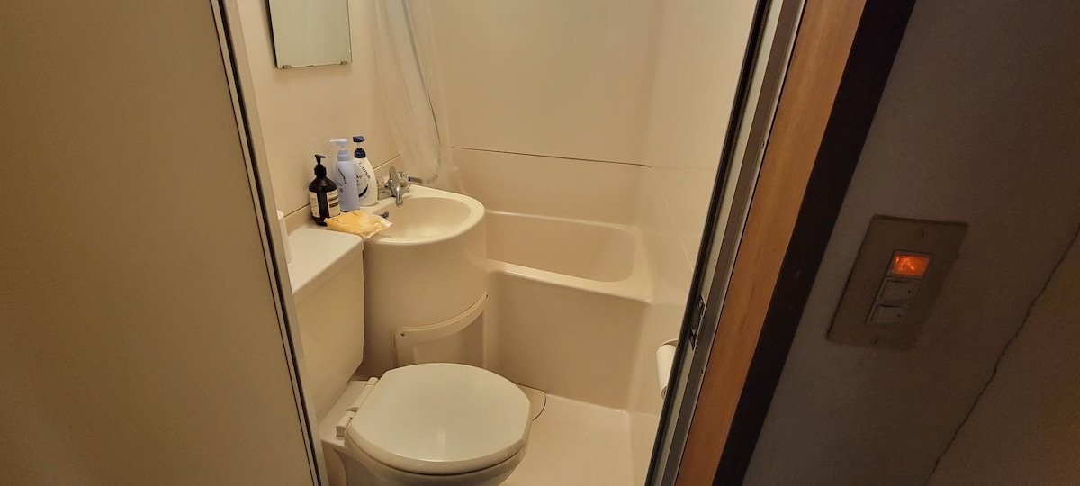 A compact bathroom features a bathtub with a shower, a toilet, and a sink. Personal care items are neatly arranged on the counter. The space is illuminated with soft lighting, creating a functional area for daily routines.