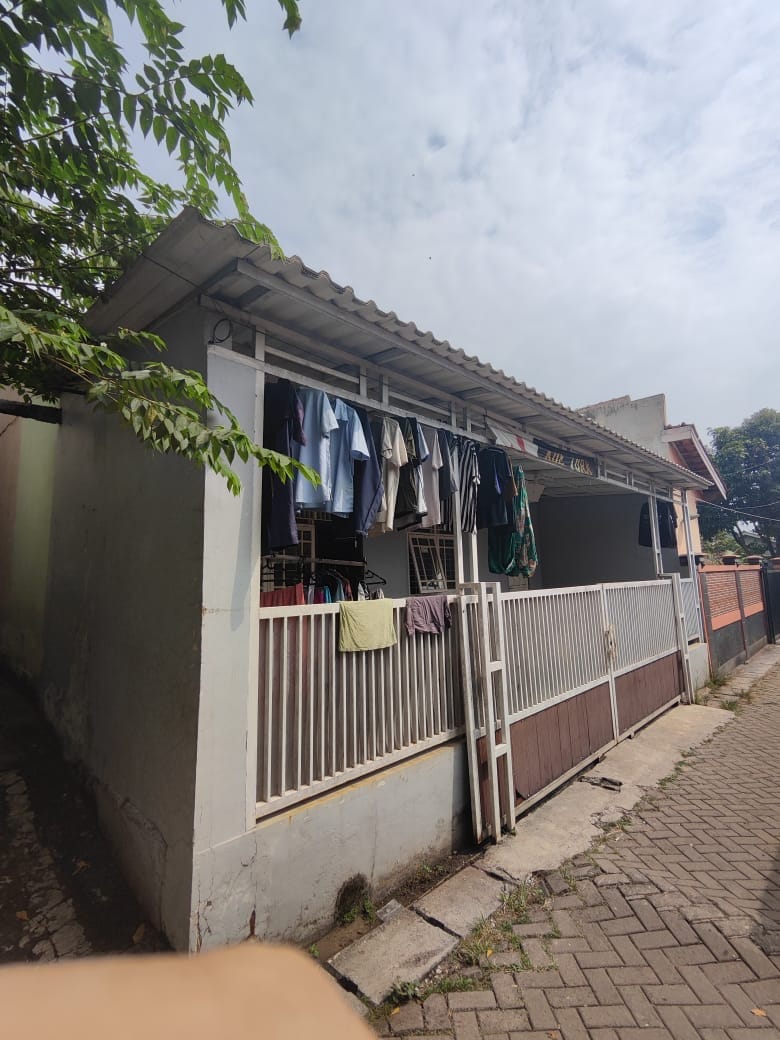 A simple home exterior is visible, featuring a covered porch where clothes are hung to dry. The wall is painted light grey, and the porch is bordered by a fence. Paved walkways line the side of the house, surrounded by greenery.