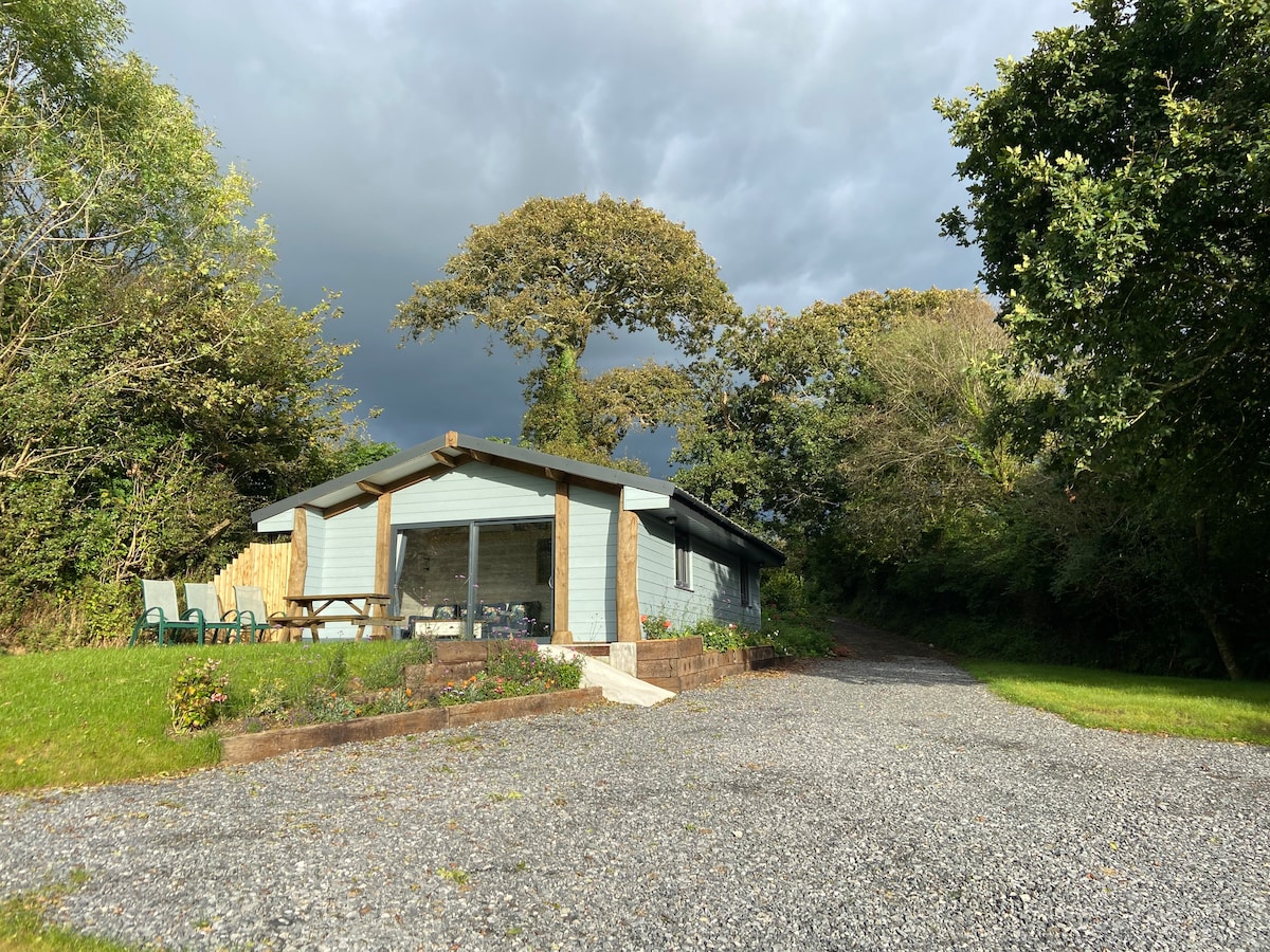 A charming lodge is positioned amidst greenery, featuring a covered entrance and a spacious gravel driveway. Outdoor seating is arranged on the patio, with trees framing the exterior under a dramatic cloud-filled sky.