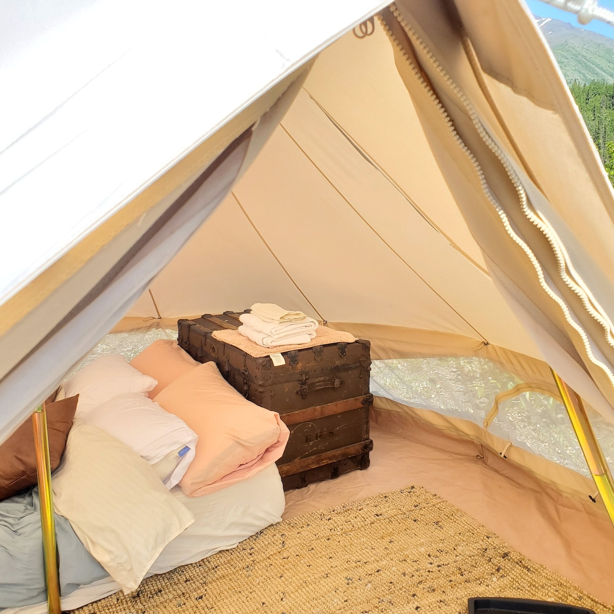 An inviting tent interior features a rustic wooden trunk at its center, surrounded by neatly arranged pillows and blankets in soft tones. Natural light floods in through the tent's windows, illuminating the earthy tones of the woven rug underfoot.