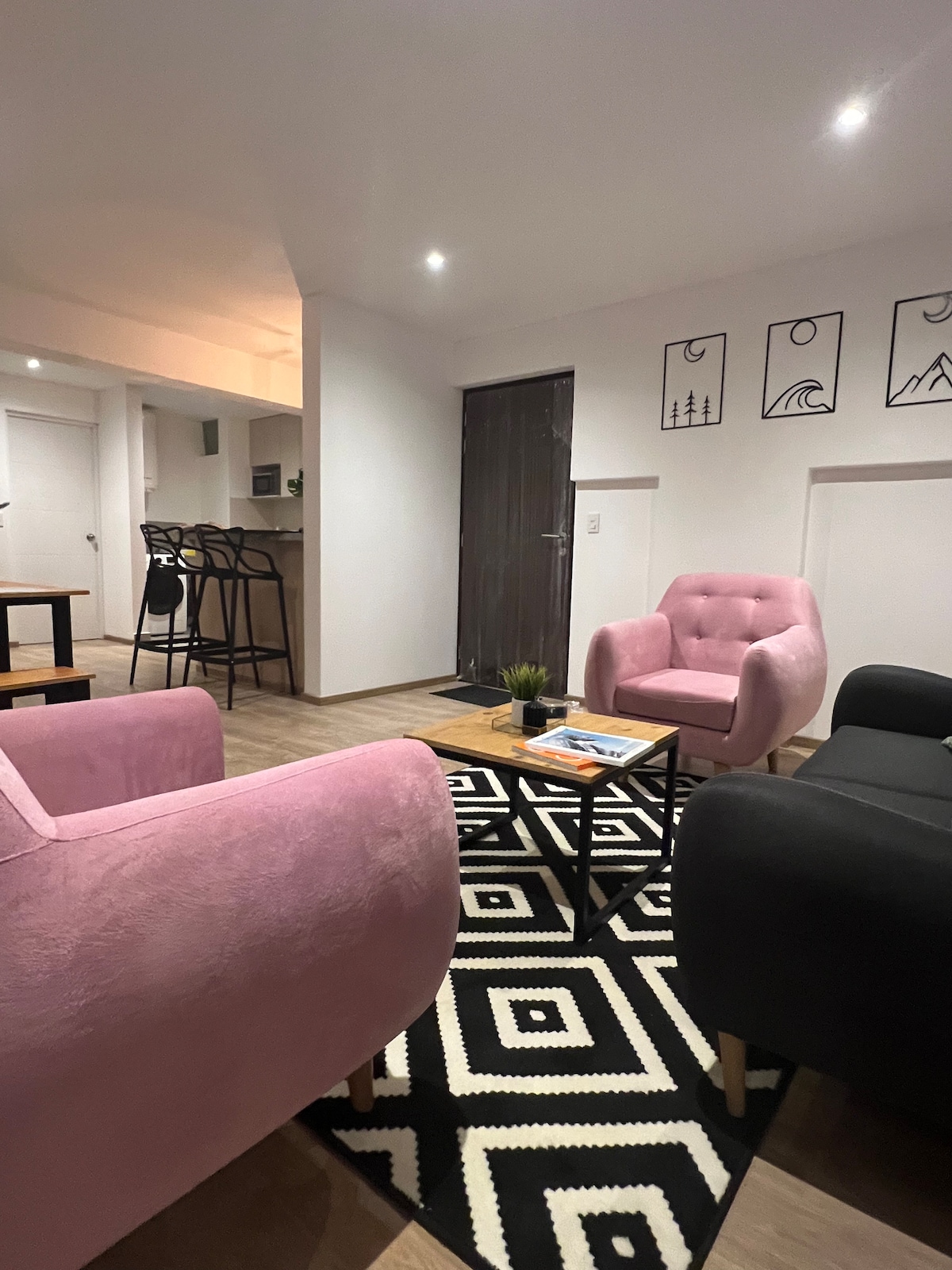 A modern living area is depicted with two pink armchairs and a black sofa arranged around a geometric-patterned rug. A small coffee table occupies the center, accompanied by a few magazines and a potted plant. The subtle lighting enhances the welcoming ambiance.