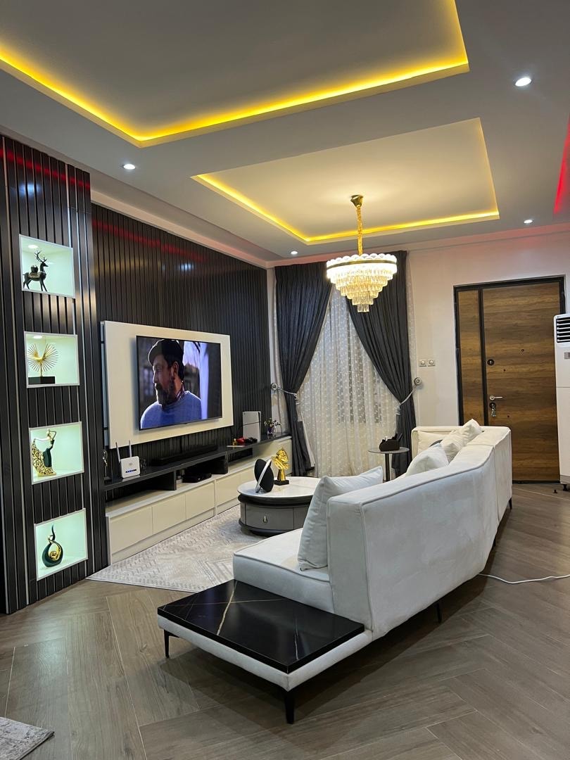 A modern living room features a sleek white sofa with black accents and a decorative side table. A large television is mounted on the wall, framed by stylish shelving displaying decor items. Elegant drapes frame the window, while recessed lighting and a chandelier add ambiance.
