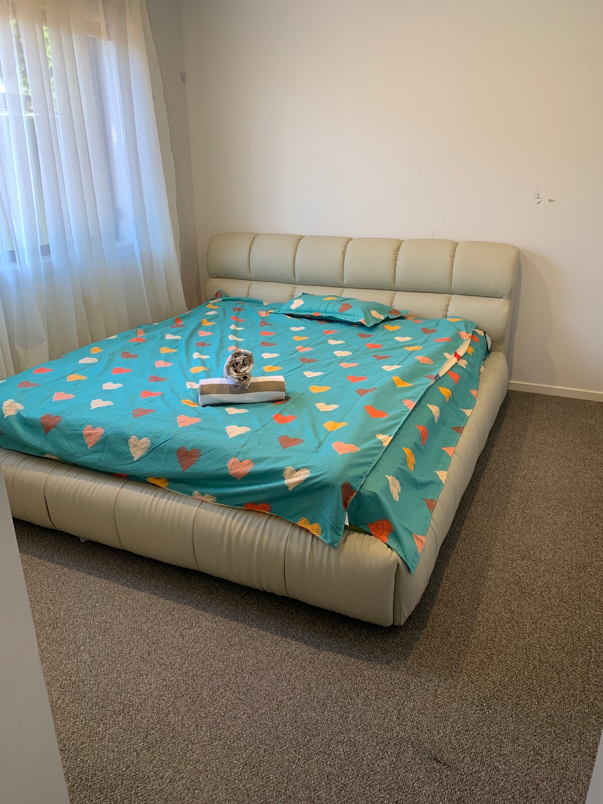 A spacious bed is centered in the room, dressed in a colorful heart-patterned coverlet. Soft light filters through sheer curtains, illuminating the neutral-colored walls and carpet. A towel is neatly placed at the head of the bed, adding a touch of hospitality.