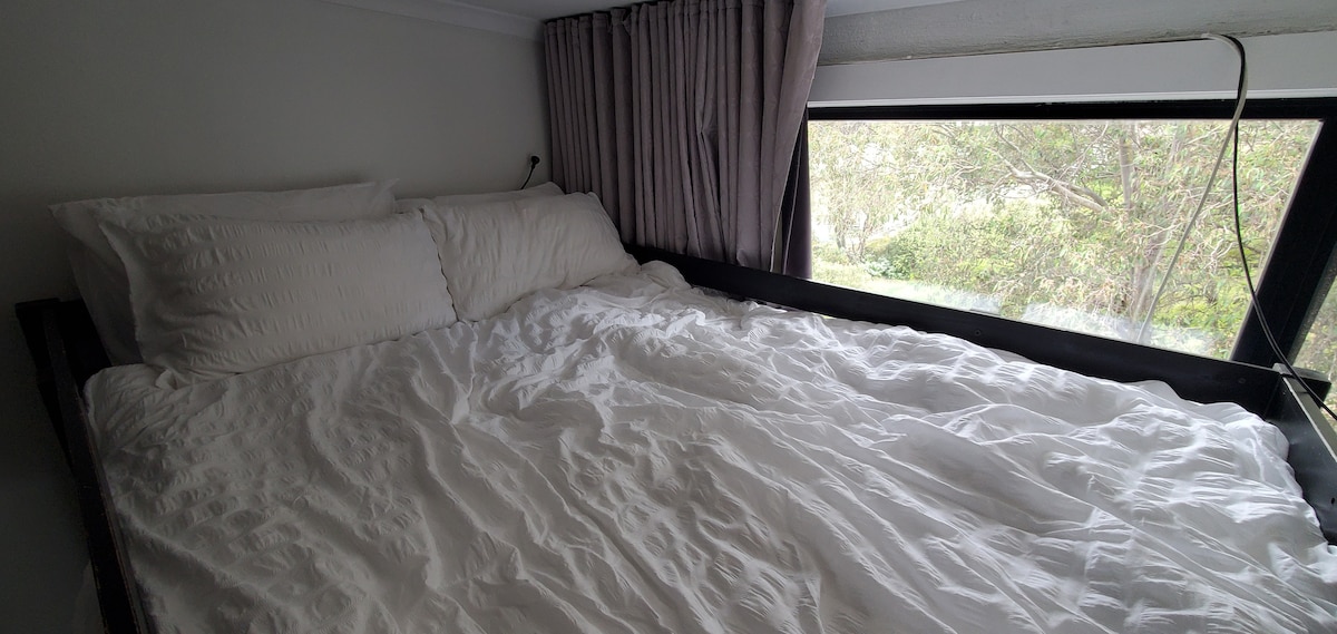 A loft bed is softly layered with white linens, providing a cozy sleeping space. Large windows allow natural light to fill the area, showcasing views of greenery outside. Curtain panels frame the windows, offering privacy when needed.