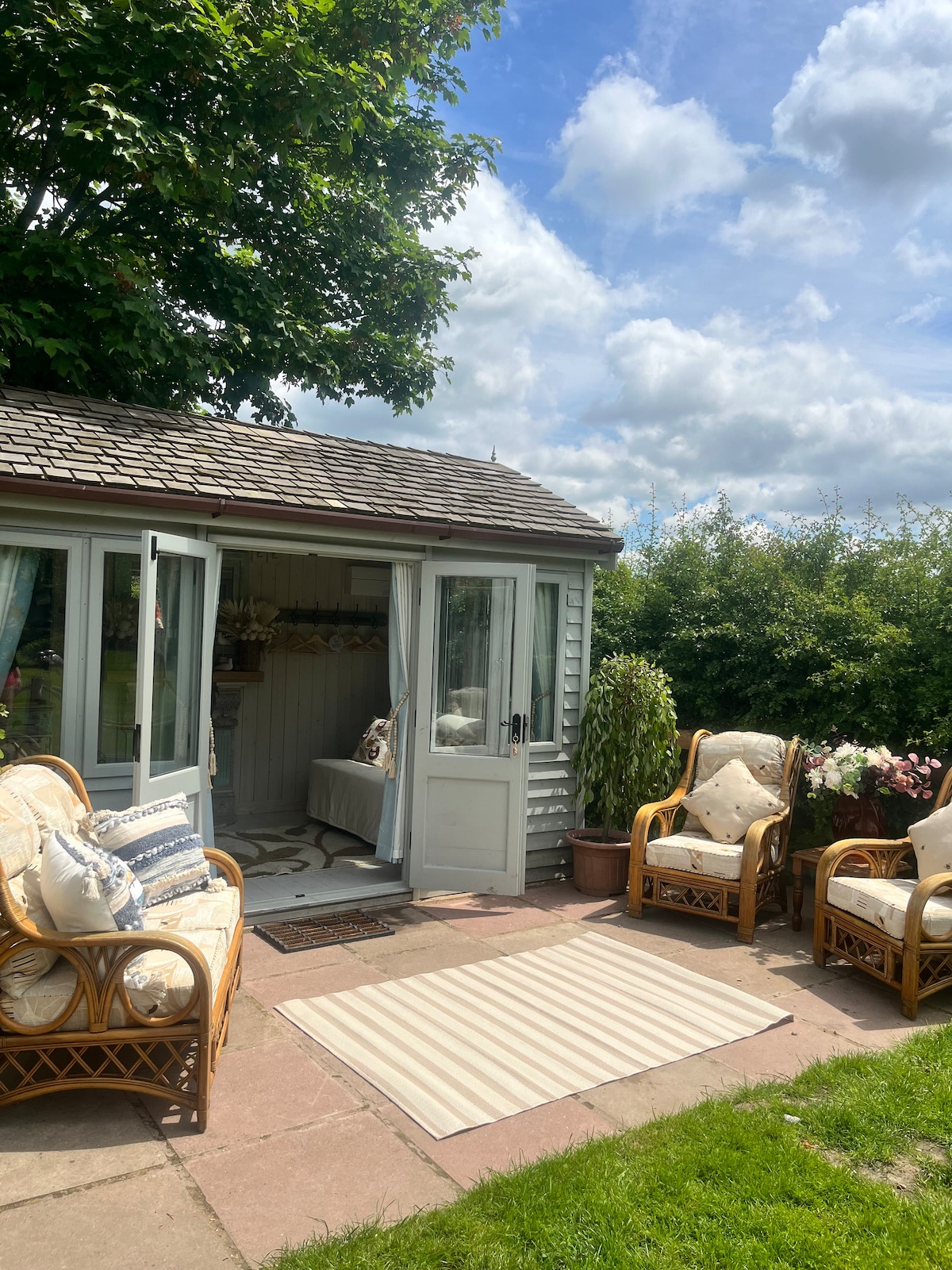 An inviting outdoor seating area is displayed in front of the cabin, featuring two wicker chairs and a small table. A light-coloured area rug lies on the patio, leading to the cabin doors, which are open, welcoming fresh air and light. Lush greenery surrounds the space.