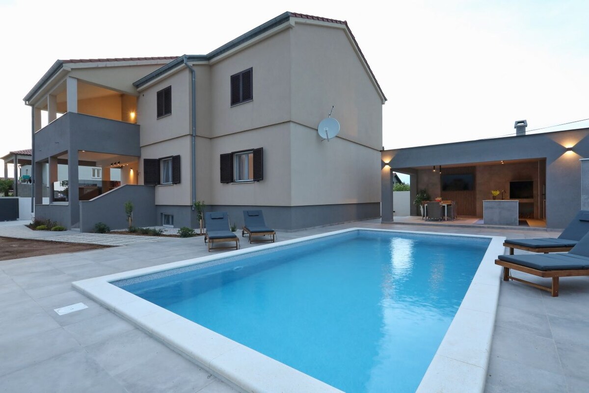 A modern villa is showcased with a private pool surrounded by a spacious terrace. Comfortable lounge chairs are positioned near the pool, while a summer kitchen and dining area are visible in the background. The building features a light-colored exterior complemented by dark window shutters.