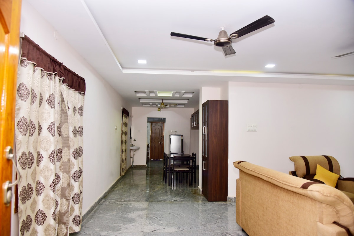 A spacious living area is shown, featuring a polished marble floor and soft lighting. A dining table is positioned near the kitchen, which is partially visible. Decorative curtains frame the windows, and a sofa sits comfortably against the wall.