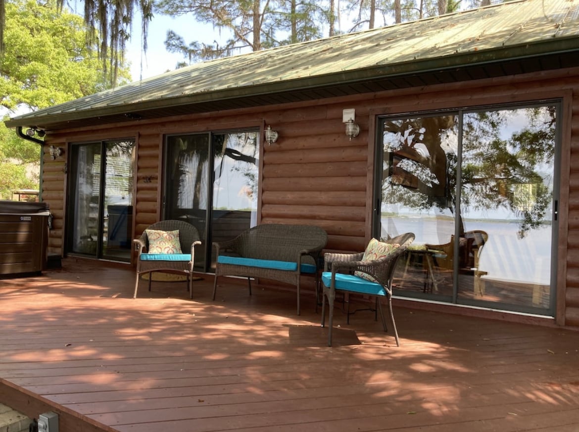 A spacious deck features comfortable seating with decorative cushions, providing a relaxing spot to enjoy views of the lakeside. Large glass doors reflect natural light and connect the outdoor space to the cabin's interior, inviting relaxation in a serene environment.