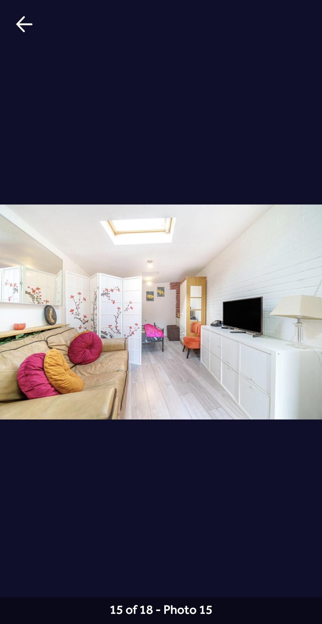A bright living area features a comfortable sofa adorned with colorful cushions. A large mirror reflects natural light, enhancing the space's openness. A television sits on a stylish sideboard, while a cozy armchair and decorative elements create a welcoming atmosphere, complemented by soft wood flooring.