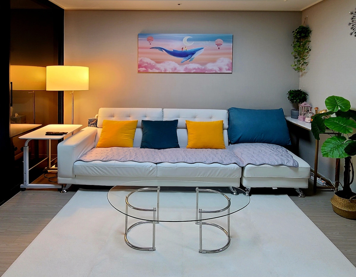 A modern living area features a bright white sofa adorned with blue and yellow cushions. A glass coffee table rests on a soft rug, while a warm lamp provides gentle lighting. Artwork depicting a whale floats above the sofa, adding a touch of whimsy.