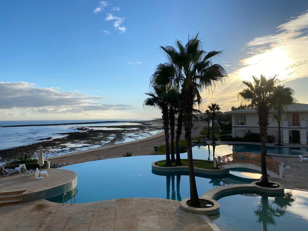 An infinity pool reflects the skies above, outlined by palm trees swaying gently in the breeze. The pool area overlooks a serene beach and calm waters, with gentle waves lapping at the shore, creating a peaceful coastal view.