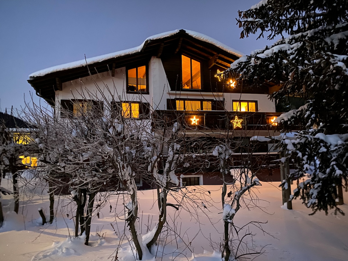 The exterior of a two-story chalet is displayed during twilight, surrounded by freshly fallen snow. Warm light spills from large windows, illuminating the balcony. Evergreen trees frame the scene, while festive decorations enhance the inviting ambiance in the winter setting.