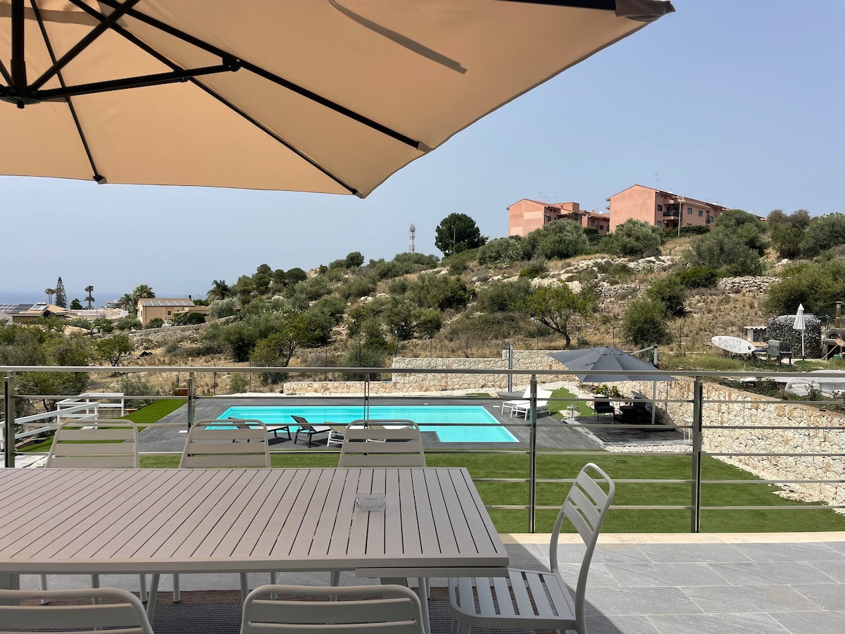 An outdoor dining area is visible under a large umbrella, featuring a long table and several chairs. The view includes a swimming pool surrounded by greenery, with distant hills and residential buildings in the background, creating a serene outdoor setting.