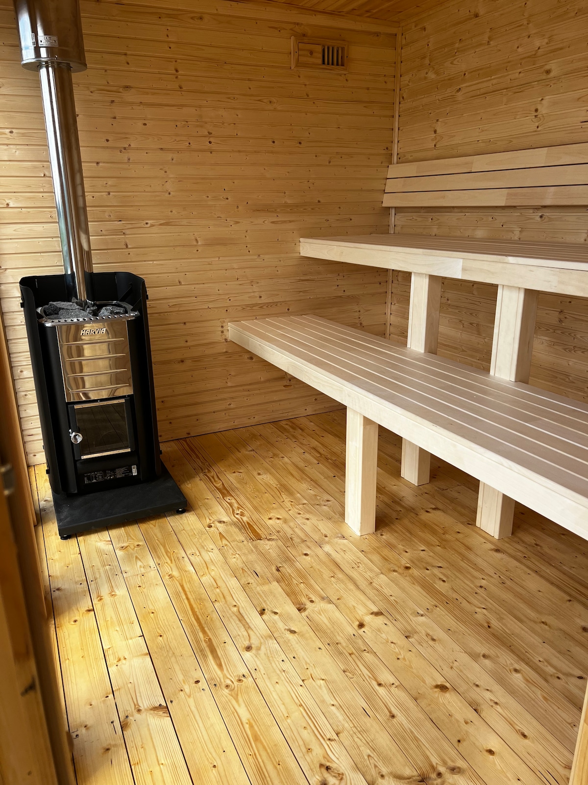 A wooden sauna interior showcases smooth pine paneling and a wooden floor. Benches line the walls, providing seating. A modern sauna stove is positioned against one wall, adding to the space's functionality. Natural light enhances the warm atmosphere within the sauna.