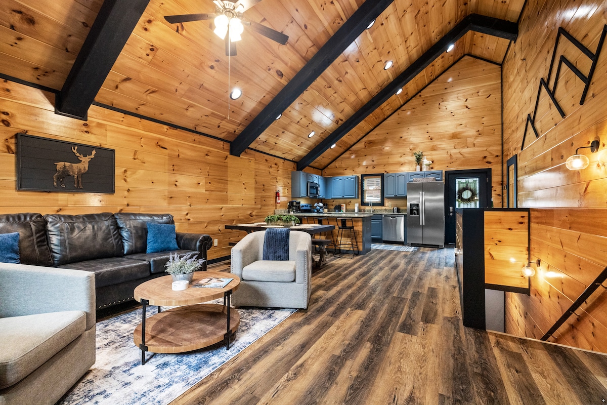 A spacious living area features wooden walls and high ceilings, creating an open atmosphere. Comfortable seating is arranged around a circular wooden coffee table, complementing the rustic decor. The kitchen area is visible in the background, showcasing modern appliances and cabinetry.