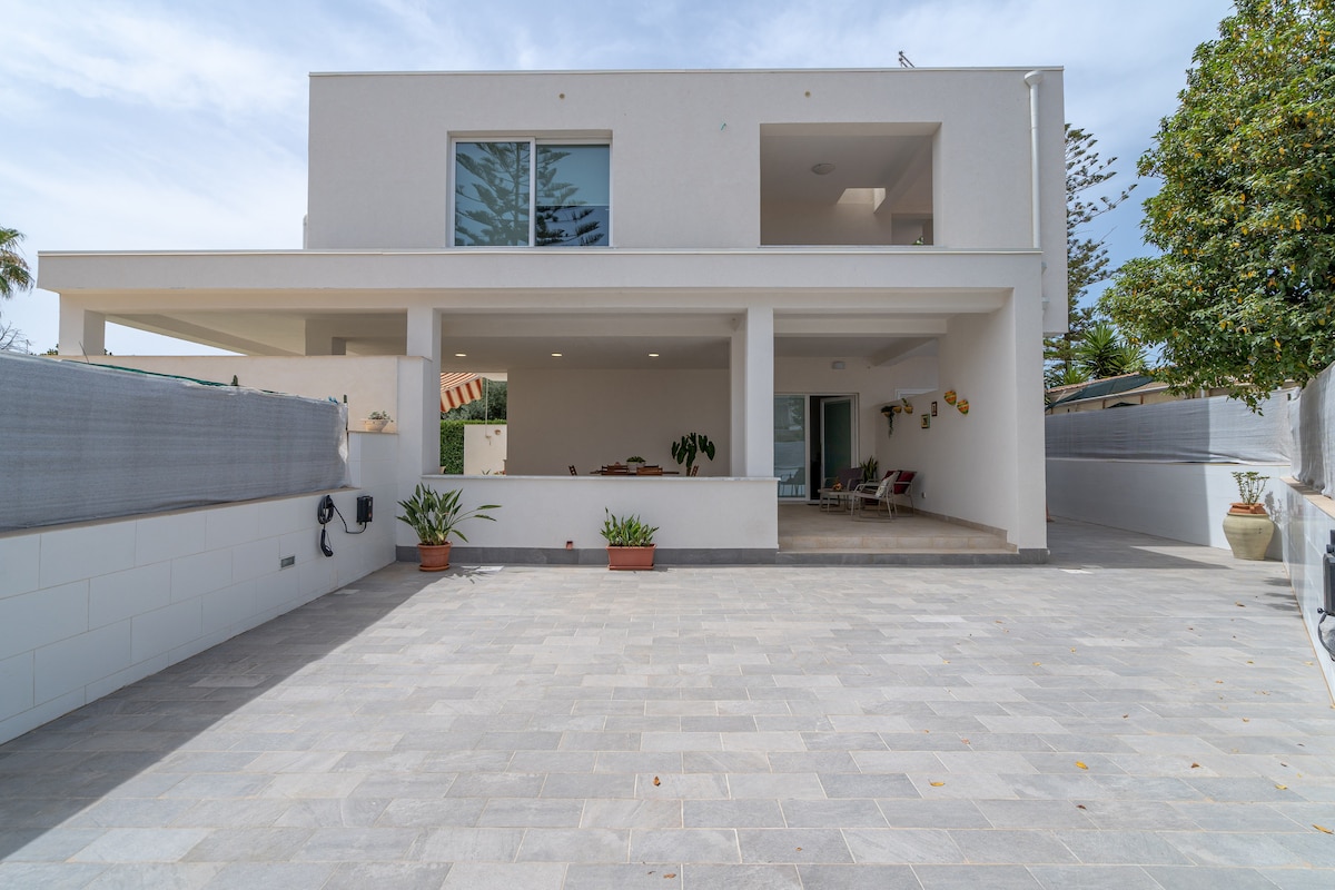 The modern villa exterior features a spacious patio area bordered by potted plants. Large glass doors lead into a bright interior, while the surrounding landscape includes greenery and an open driveway paved with stone tiles.