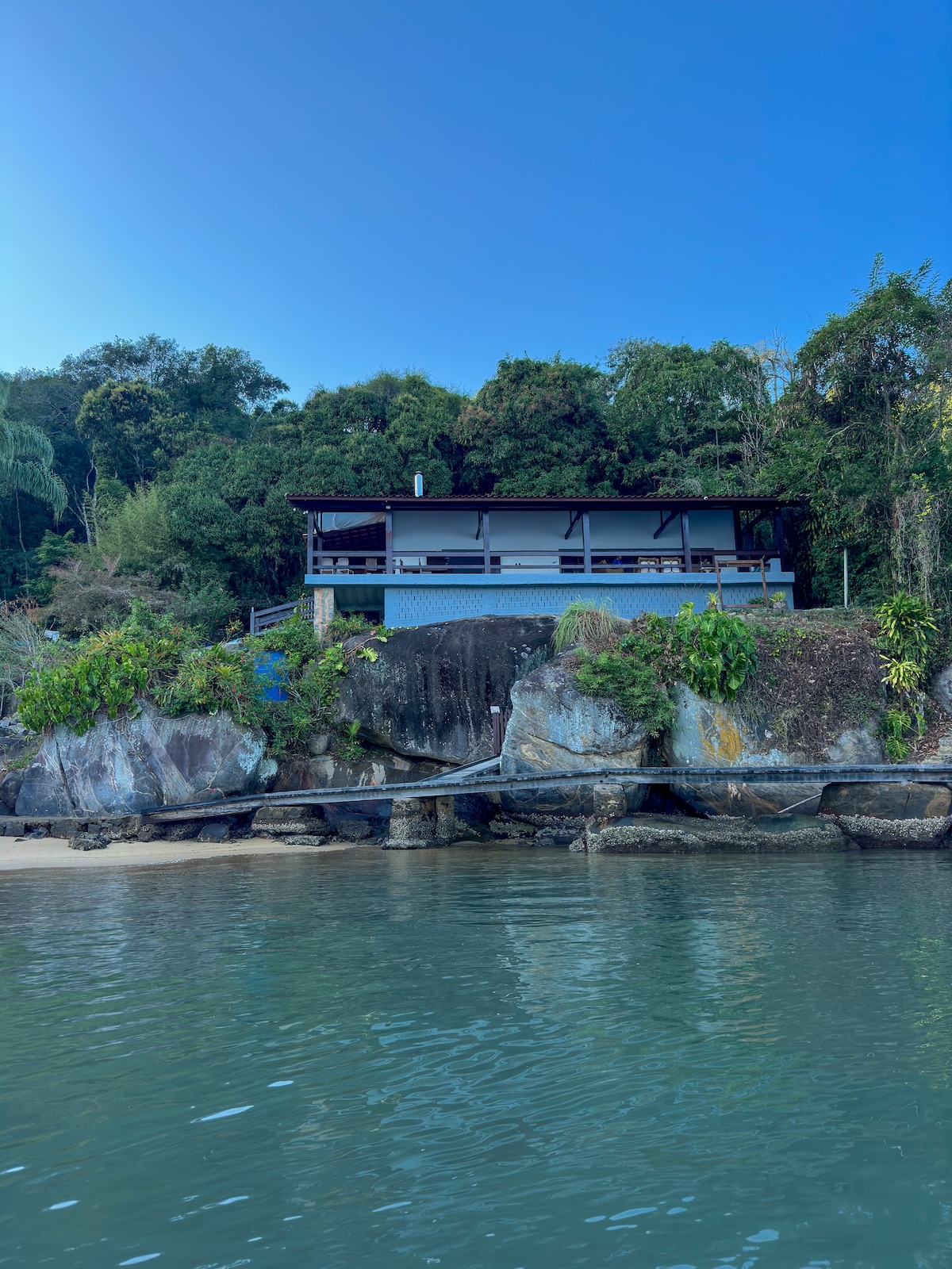 A modern two-story house rests on a rocky outcrop, surrounded by lush greenery. The property features a spacious deck, offering views of the sea. The clear water reflects the blue sky, complementing the coastal landscape.