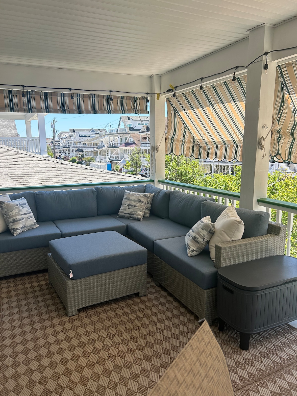 A large, comfortable seating area features a sectional sofa adorned with decorative pillows. A small side table complements the seating arrangement. The porch is shaded by striped awnings, and a patterned rug adds texture to the space, surrounded by views of neighboring houses.