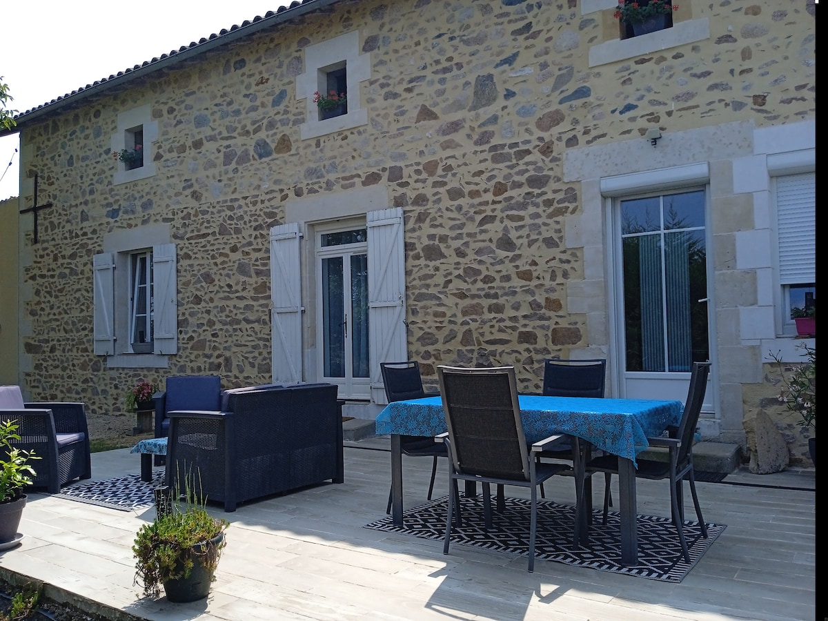A spacious outdoor area is highlighted by a stone facade of a house. A large dining table with chairs sits on a wooden patio, complemented by potted plants. Light blue tablecloth adds a pop of color, and potted flowers adorn the textured walls and window ledges.