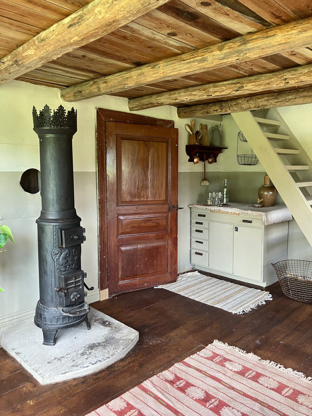 A traditional cast iron stove is positioned beside a wooden door, contributing to the rustic charm of the interior. A staircase leads to the upper level, with a small work area visible in the background. The wooden ceiling beams and woven rugs add warmth to the space.
