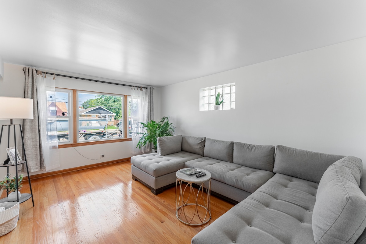 A modern living room features a spacious gray sectional sofa paired with a circular metal coffee table. Bright light enters through a large window, accompanied by a smaller glass block window. A vibrant plant adds a touch of greenery, enhancing the simple decor.