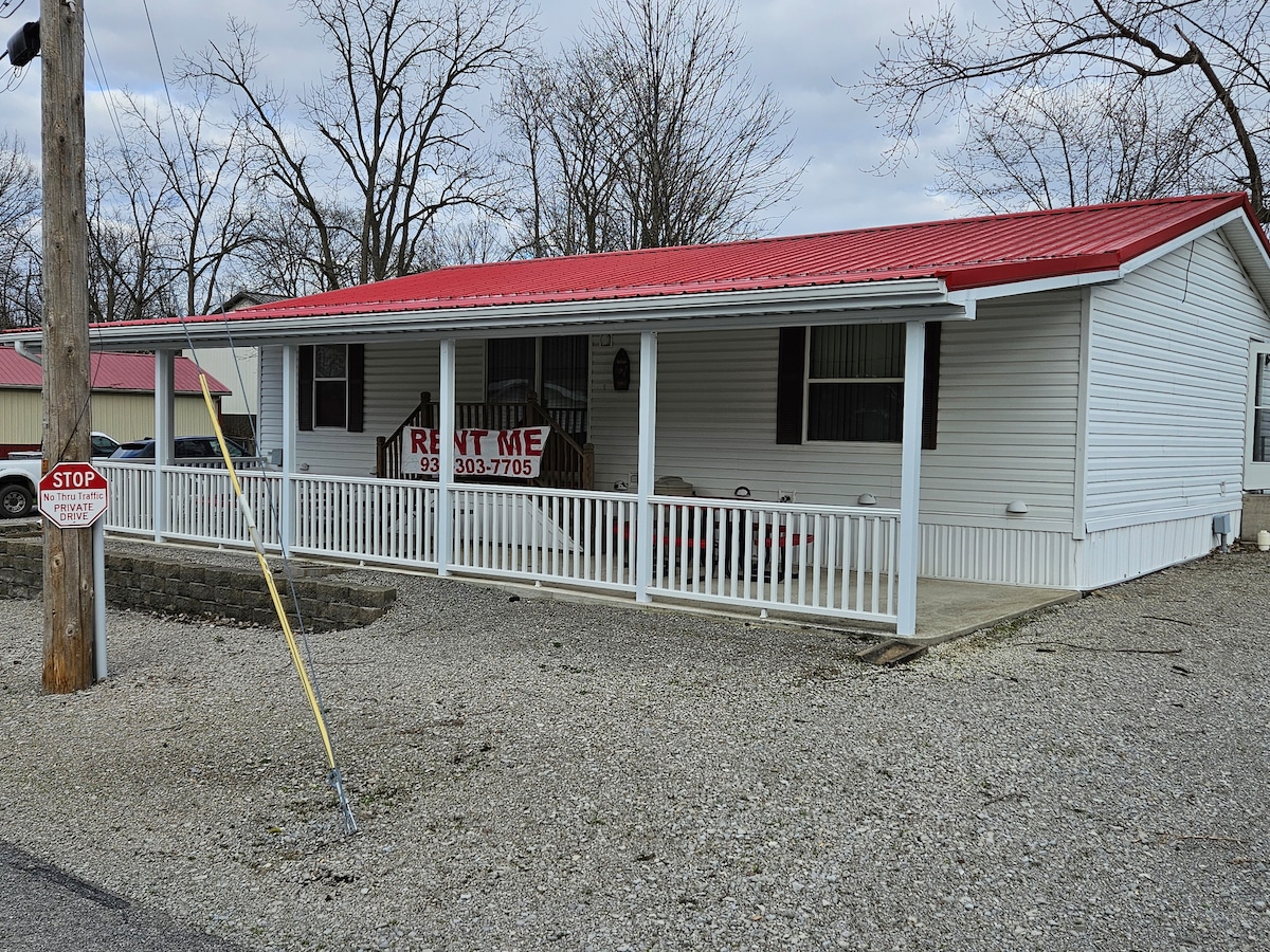 A single-story house is displayed with a red metal roof and white exterior. A welcoming front porch features a railing and a sign indicating availability for rent. The surrounding area includes gravel and a stop sign, with bare trees in the background.