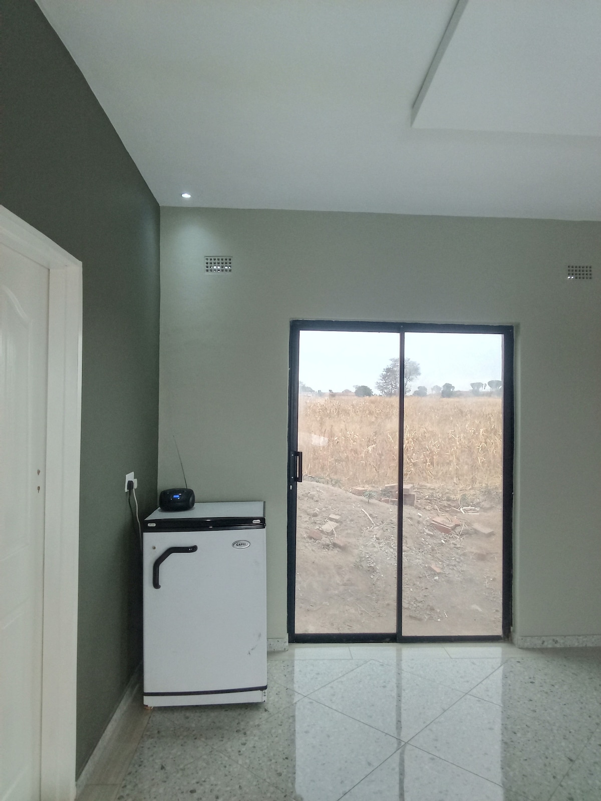 A compact kitchen area is visible, featuring a small refrigerator beside a doorway. A sliding glass door allows natural light to enter and provides a view of the outdoor landscape, which includes open fields. The interior walls are painted in a muted green shade.