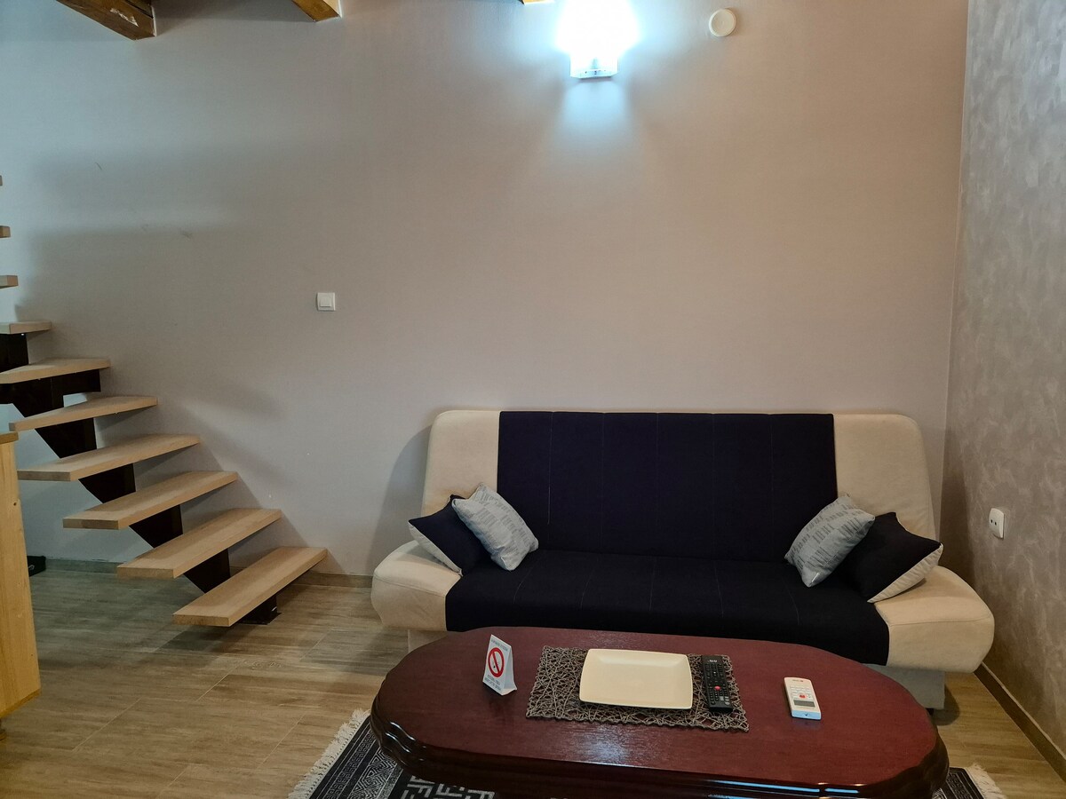 A modest living area is presented with a two-toned sofa, paired with decorative pillows. A wooden coffee table sits in front, complemented by a remote control and a decorative tray. Natural light illuminates the space, enhancing the warm tones of the wooden staircase in the background.