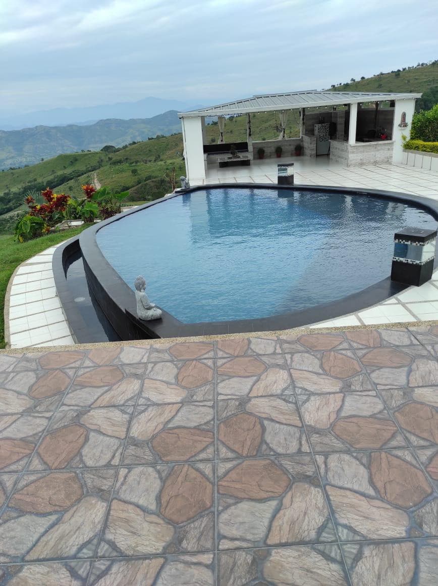 An infinity pool offers a panoramic view of the surrounding hills and valleys. The pool's smooth surface reflects the sky, while a well-maintained deck features geometric stone patterns. A covered outdoor kitchen area is visible in the background, surrounded by lush greenery.