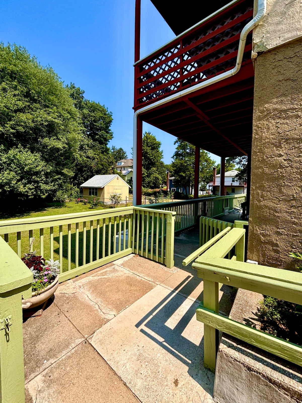 A welcoming entrance path is bordered by a green railing, leading toward a patio area under a covered balcony. Lush greenery is visible in the surrounding yard, contributing to a serene environment. The walkway is shaded, providing a comfortable transition into the apartment.