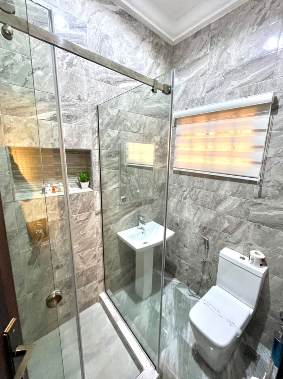 A modern bathroom is featured, showcasing a glass-enclosed shower with elegant gray tiling. A compact vanity is positioned beside a sleek toilet. Natural light is provided through a window covered with a blind, and a small plant adds a touch of greenery.