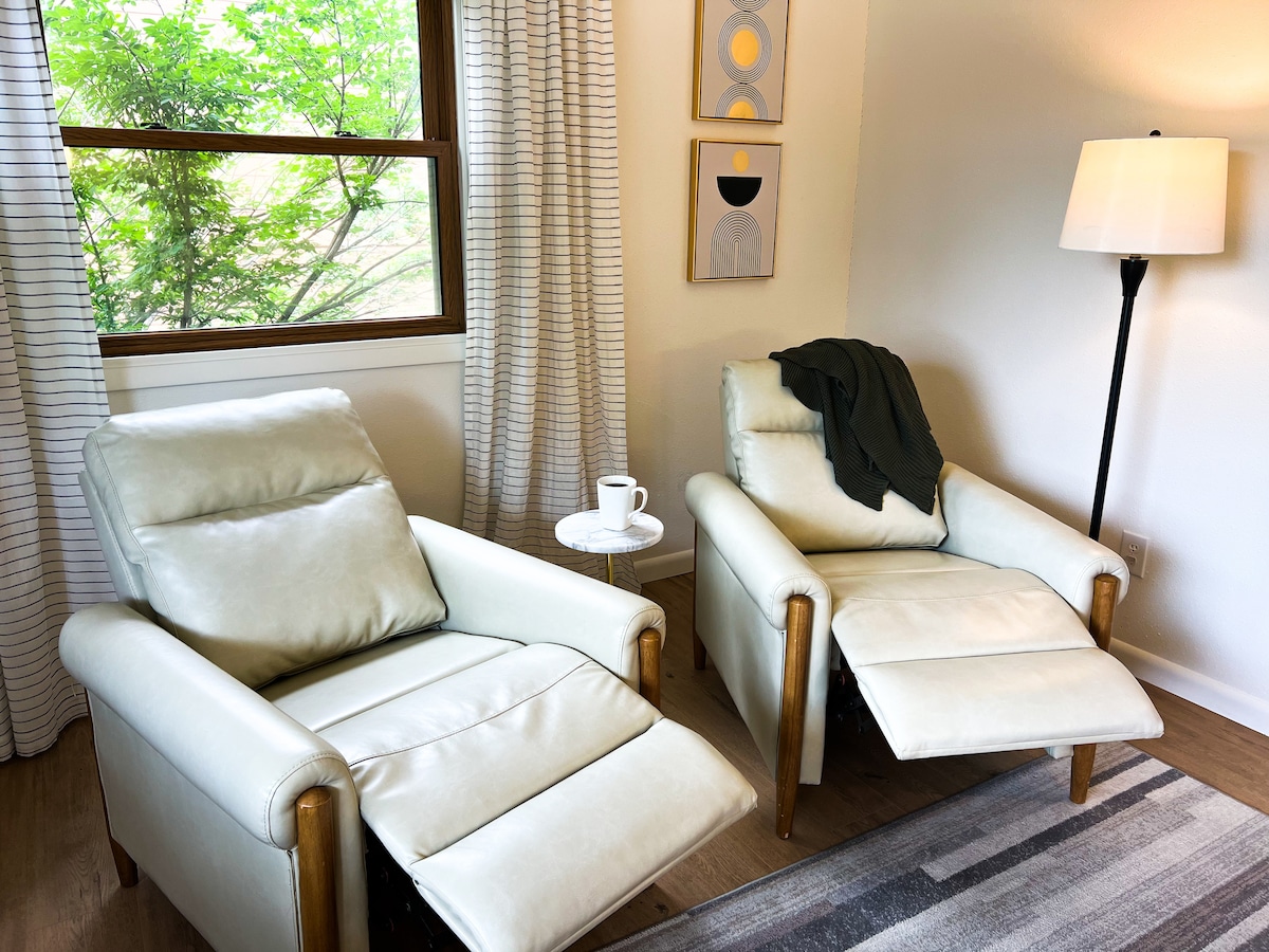 Two reclining chairs sit facing a window, allowing natural light to fill the space. A small side table holds a cup, and a light-colored throw is draped over one chair. Soft curtains frame the window, enhancing the peaceful environment.