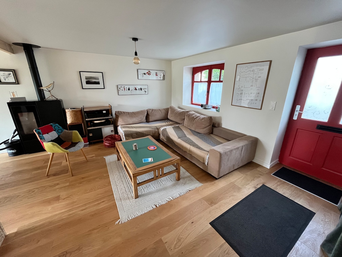 A bright living area is showcased, featuring a corner sofa arranged with a striped blanket. A low coffee table is centered on a light-colored rug. A colorful chair complements a wood-stove in the corner, while large windows provide natural light and a view of the entryway.
