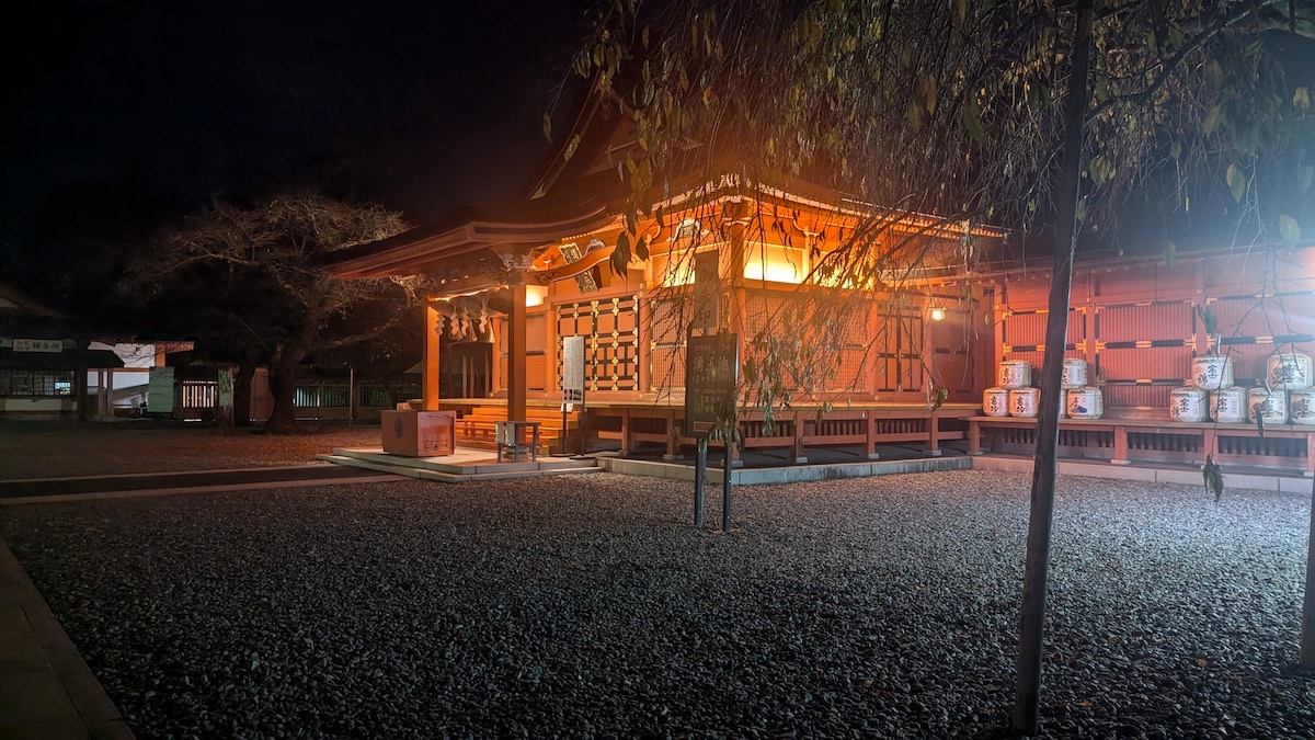 A traditional Japanese shrine is illuminated at night with warm orange lights. The structure features intricate wooden details and sliding doors, surrounded by a gravel courtyard. Lanterns and a few leafing trees add to the serene atmosphere.