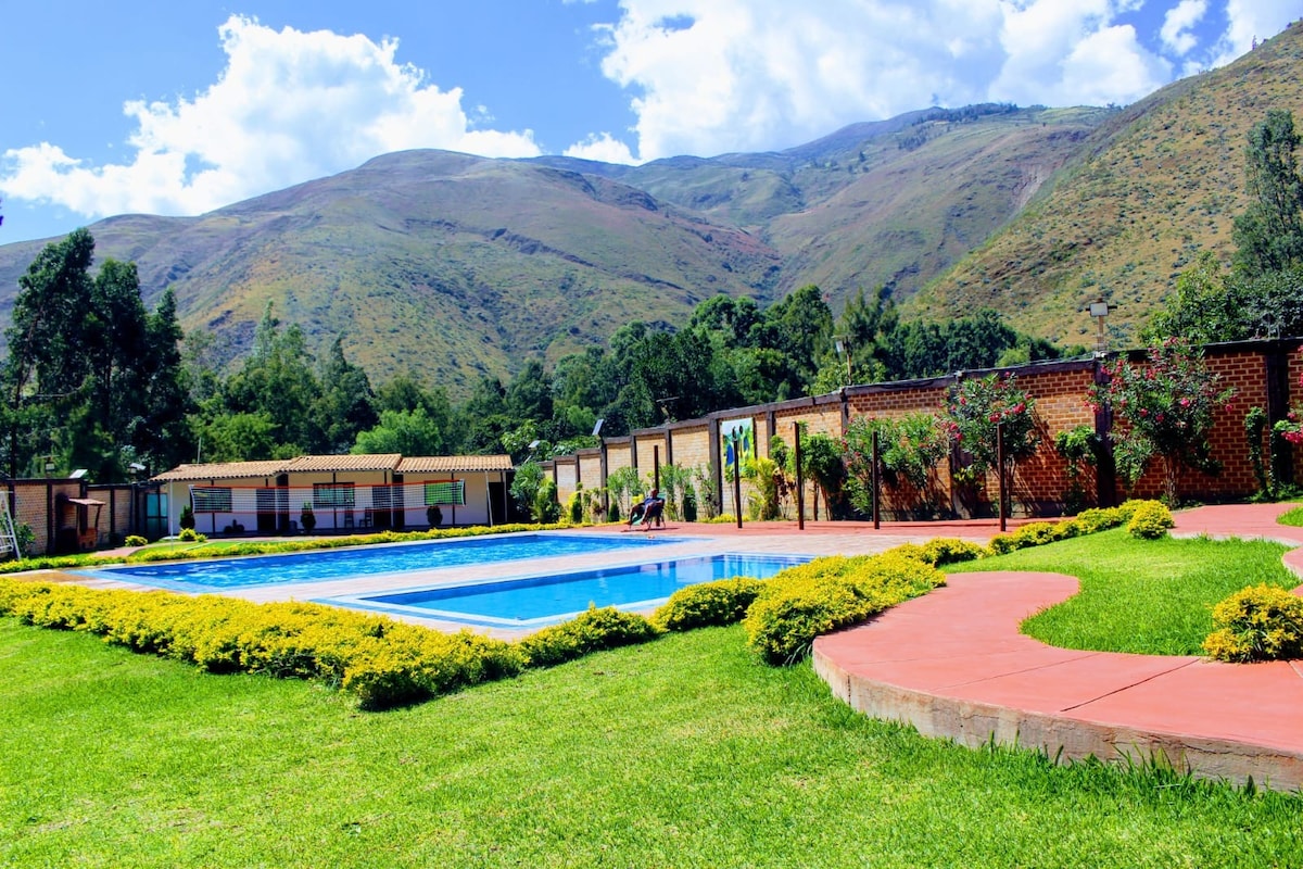 A clear view of a swimming pool surrounded by well-maintained greenery, flowers, and shrubbery. The backdrop features majestic mountains under a blue sky, with several bungalows visible in the distance, emphasizing the spacious outdoor area for relaxation.