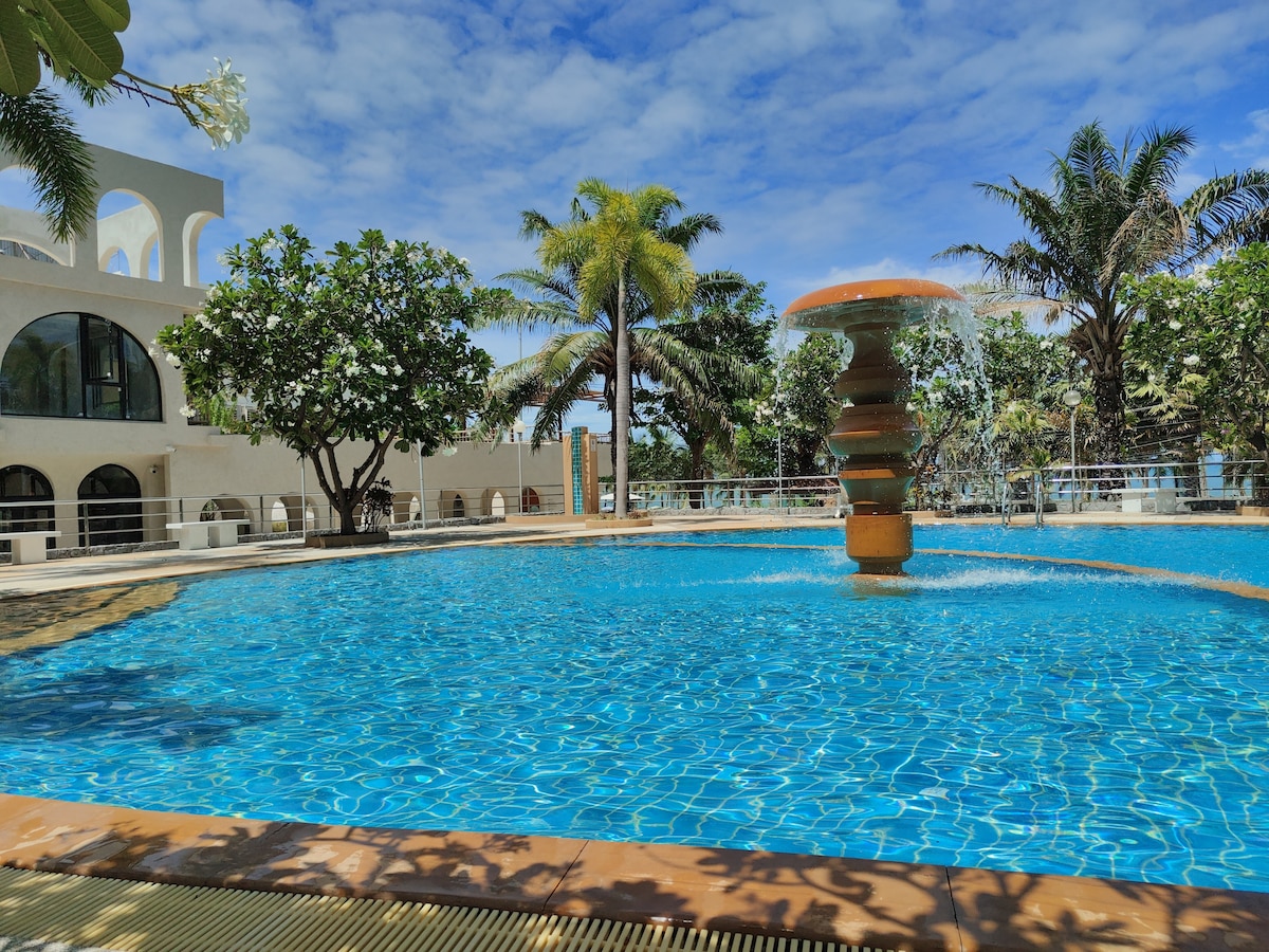 A tranquil pool area is surrounded by lush greenery and palm trees, featuring a central fountain. Crystal-clear water reflects the blue sky, and a spacious deck offers ample room for lounging. The backdrop includes a distinctive building with large arches and multiple outdoor seating sections.