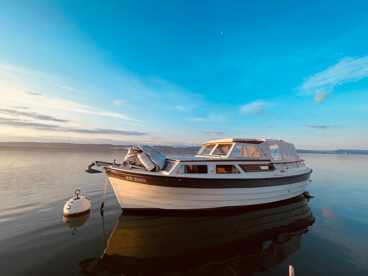 A motorboat is anchored at a buoy in the calm waters, with the sky displaying soft hues of blue. The boat's sleek exterior reveals windows that reflect the serene landscape, and a covered cockpit area suggests comfortable outdoor seating.