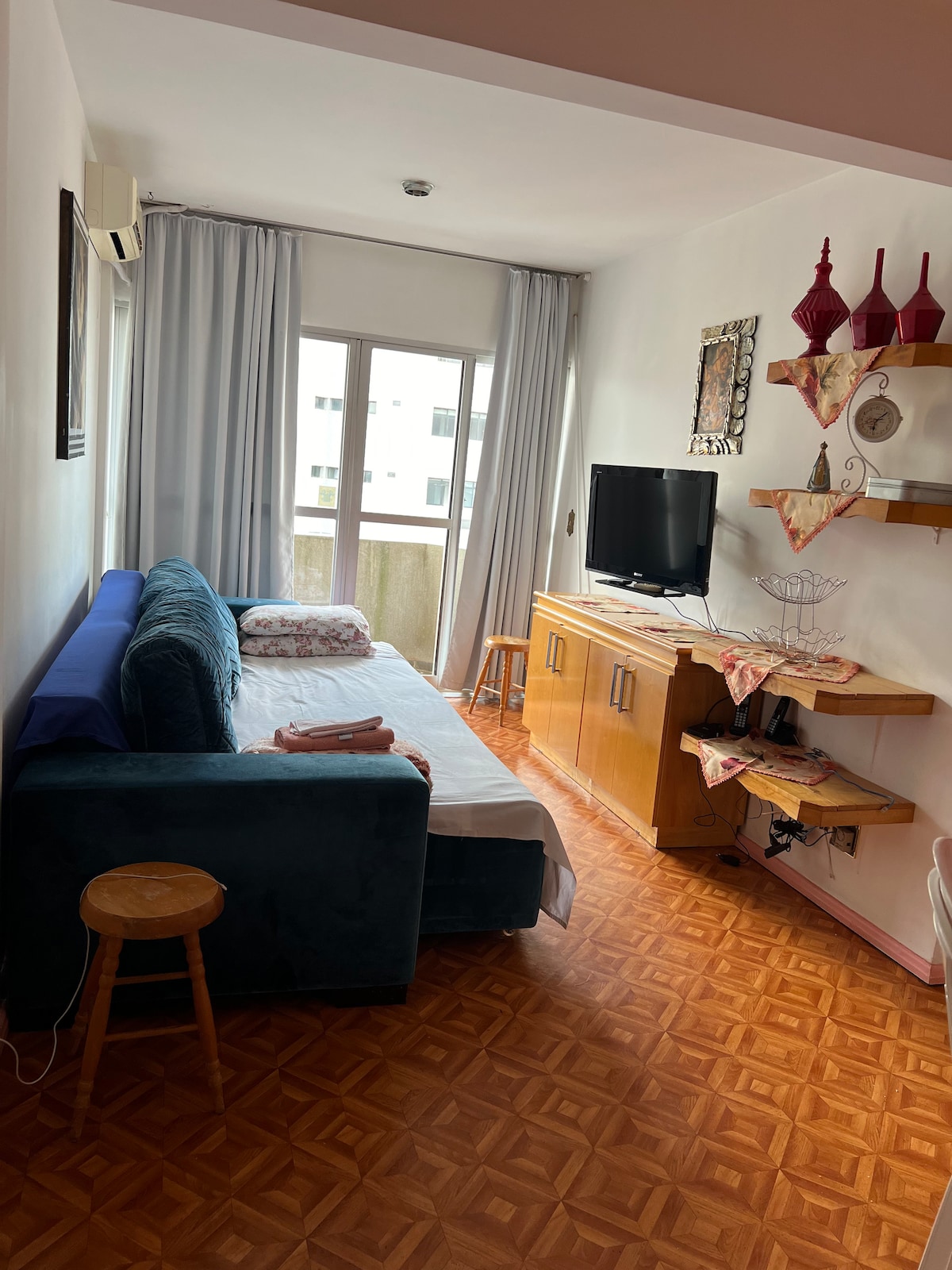 A cozy living area features a comfortable blue sofa and a small wooden stool. Natural light streams in through a large window with sheer curtains. A wooden cabinet holds a television, while a shelf displays decorative items, creating a simple yet functional space.