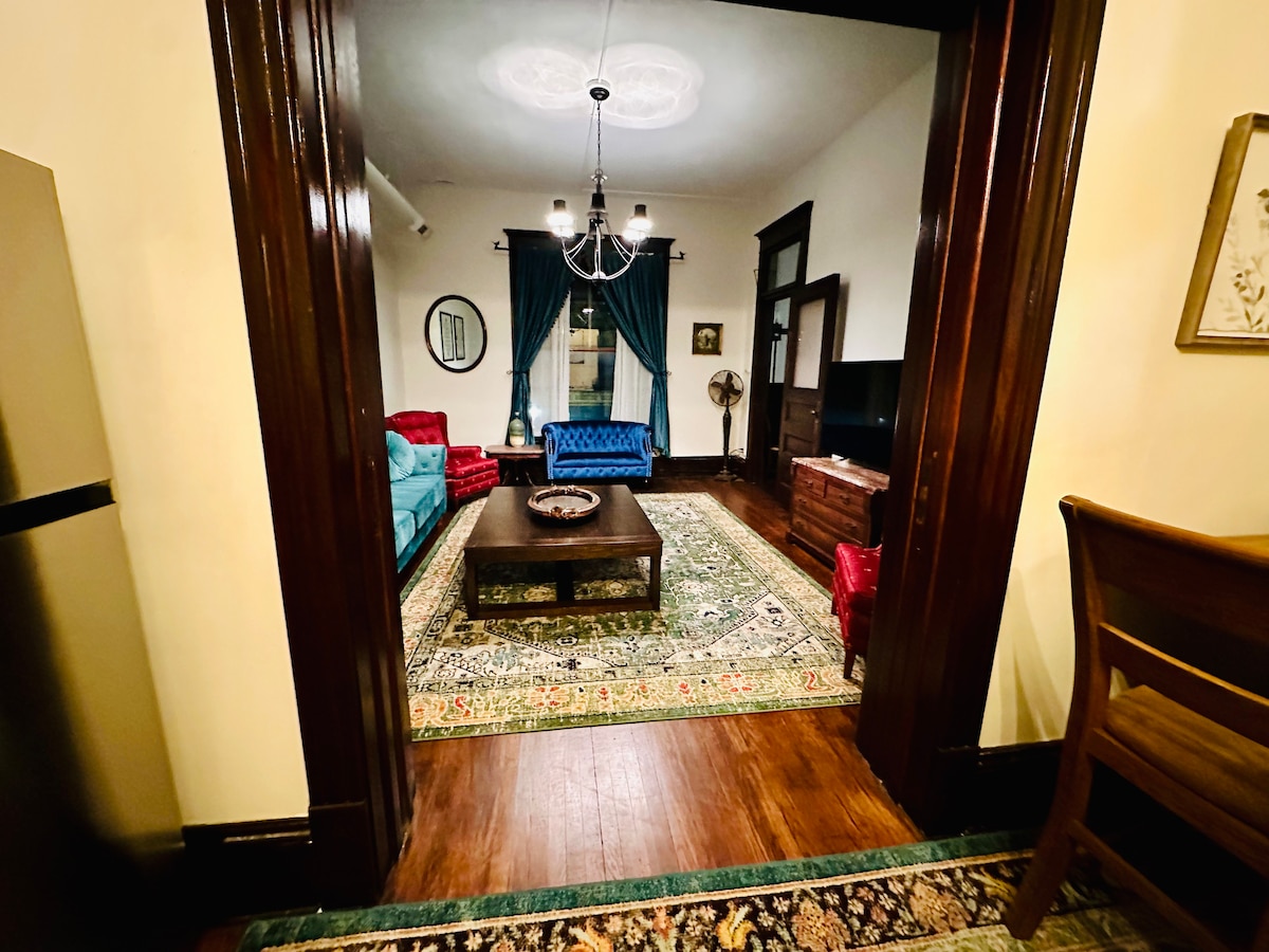 A spacious living area is showcased, featuring a blue velvet sofa and a red chair set around a central coffee table. A decorative area rug adds color, while a chandelier provides overhead illumination. Dark wooden frames complement the elegant design, with a TV positioned on a cabinet.