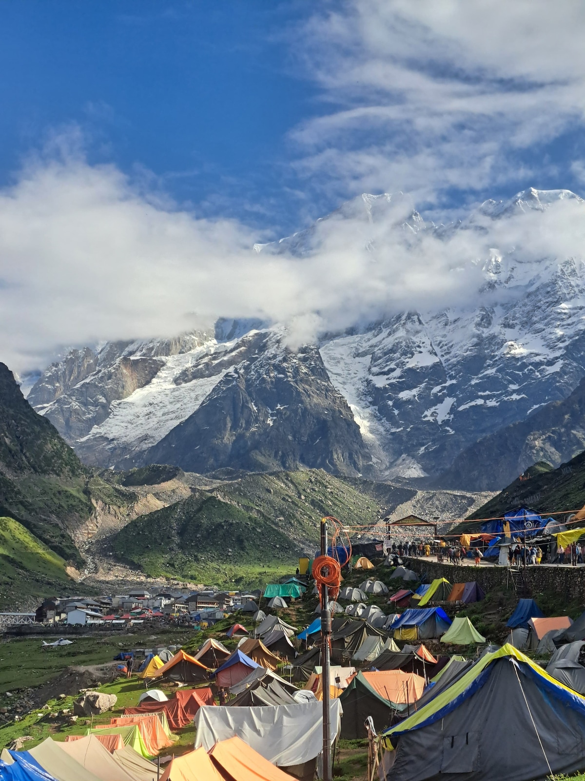 A vibrant array of colorful tents is set against a dramatic backdrop of snow-capped mountains and lush greenery. The landscape is dotted with rocky terrain and a winding path leading up the hills, creating a harmonious blend of nature and outdoor accommodation.