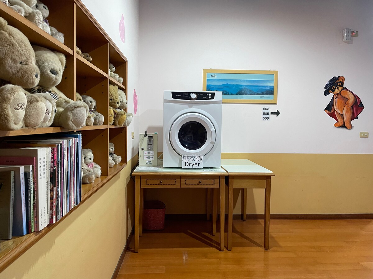 A laundry area is depicted, featuring a white dryer prominently displayed on a table. A collection of soft toys is arranged on a nearby shelf, with books neatly stacked below them. Calm wall colors and an artwork piece contribute to the relaxed ambiance.
